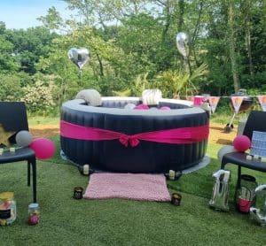 Spa gonflable décoré avec un ruban rose, ballons et accessoires dans un jardin pour un anniversaire.